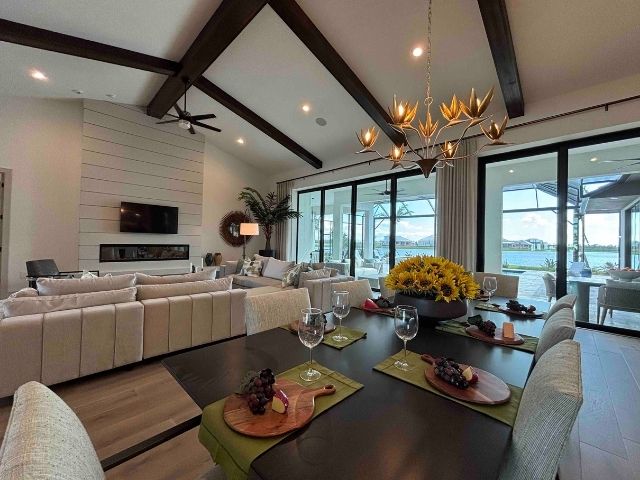photo shows the inside living area of a custom home build by neal signaature homes in wellen park. Looking from the formally dressed dining table which hosts a tray of sunflowers alongside the round charcuterie boards, the large lounge sofa faces the entertinment system, with glass patio doors to the right. Dark wood ceiling beams create a cosy feel while the low hanging artisan light offers elegant focal features. 