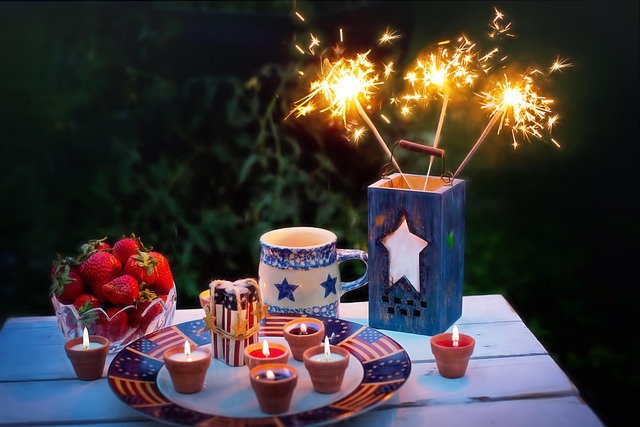 its evening, a white wooden table contrasts the dark sky. On it sits a bowl of rspberries, a blue ceramic candle holder with three handheld sparklers in it, celebrating fourth of july