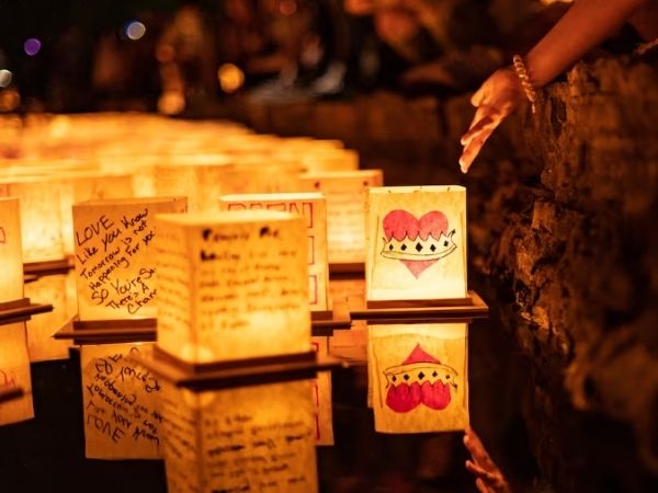lots of water lanterns floating on the water with heartfelt messages written on them. the sky is dark, the water shimmering with lights, and a hand reaches down to push the lanterns out. 