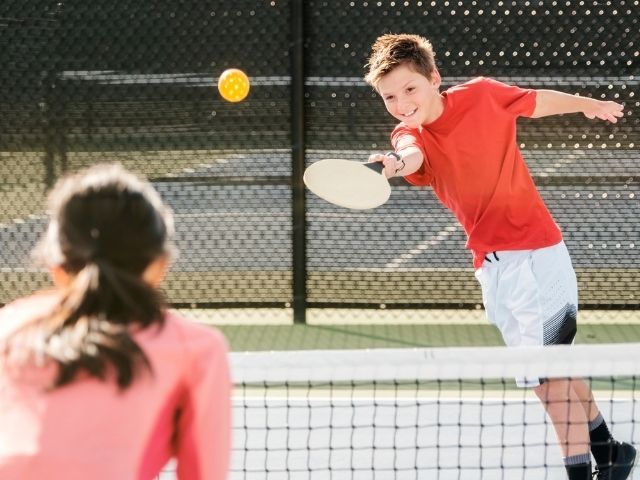a pre-teen boy in a red t-shirt serves a pickleball to a girl in a pink top. 