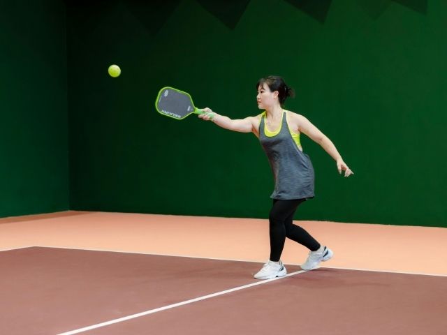 an asian women in black leggings, grey vest, and white sneakers learns how to  play pickleball at an indoor court
