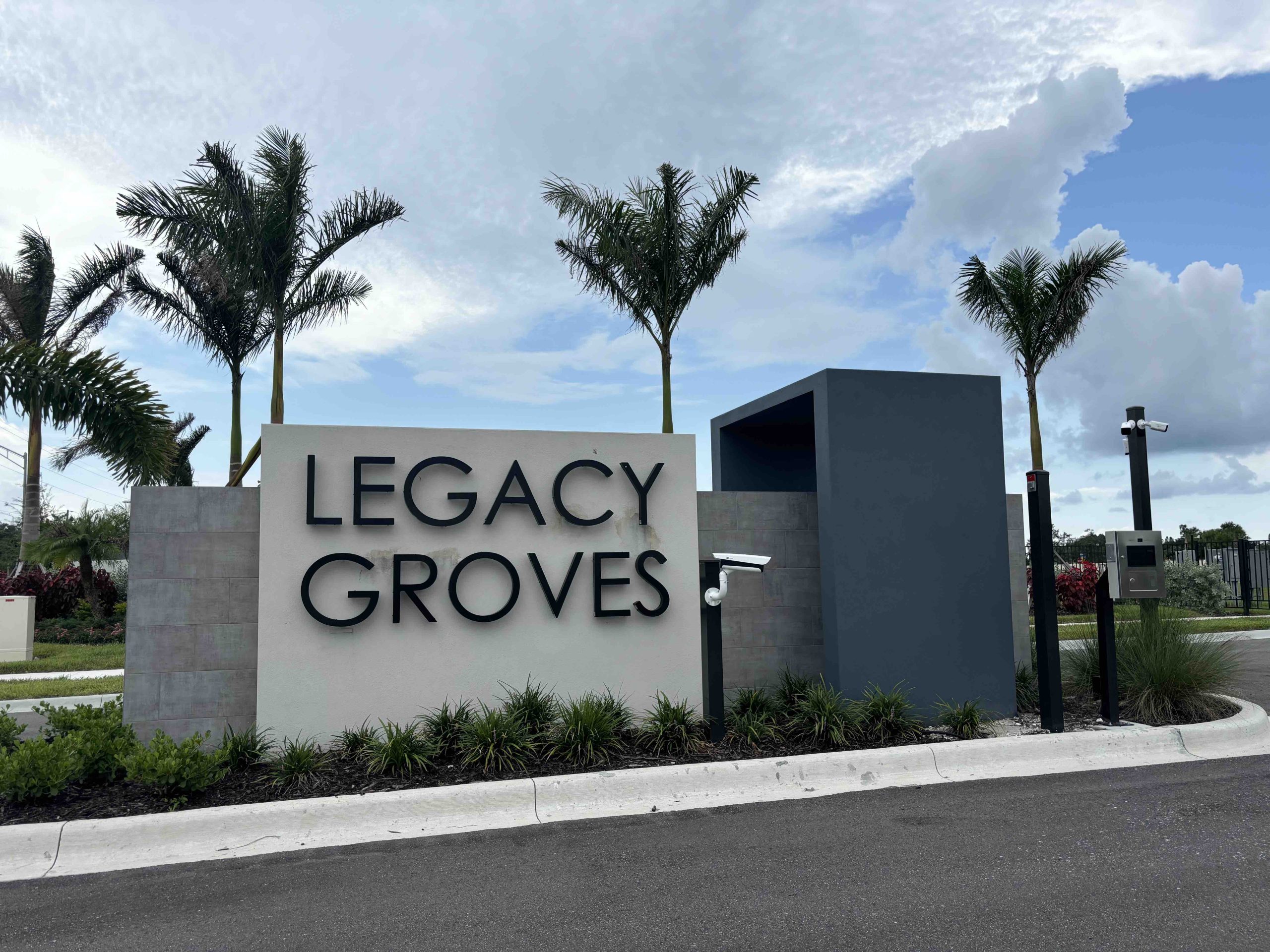 large grey stone sign at the side of the road with "legacy groves" on it, surrounded by palm trees