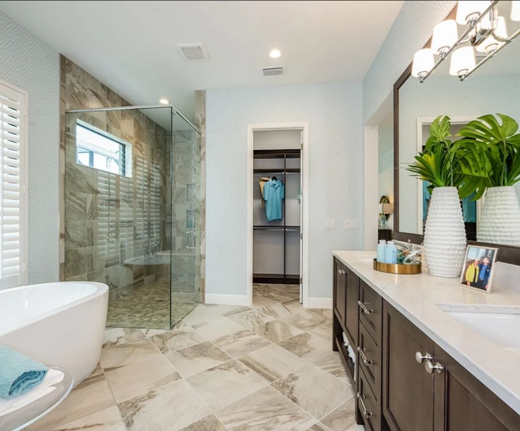 contemporary bathroom with marble floor, large freestanding bath, glass panel walk-in shower, and built in cupboards. lights shine above the mounted wall mirrors, and a plant sits on the counter top.