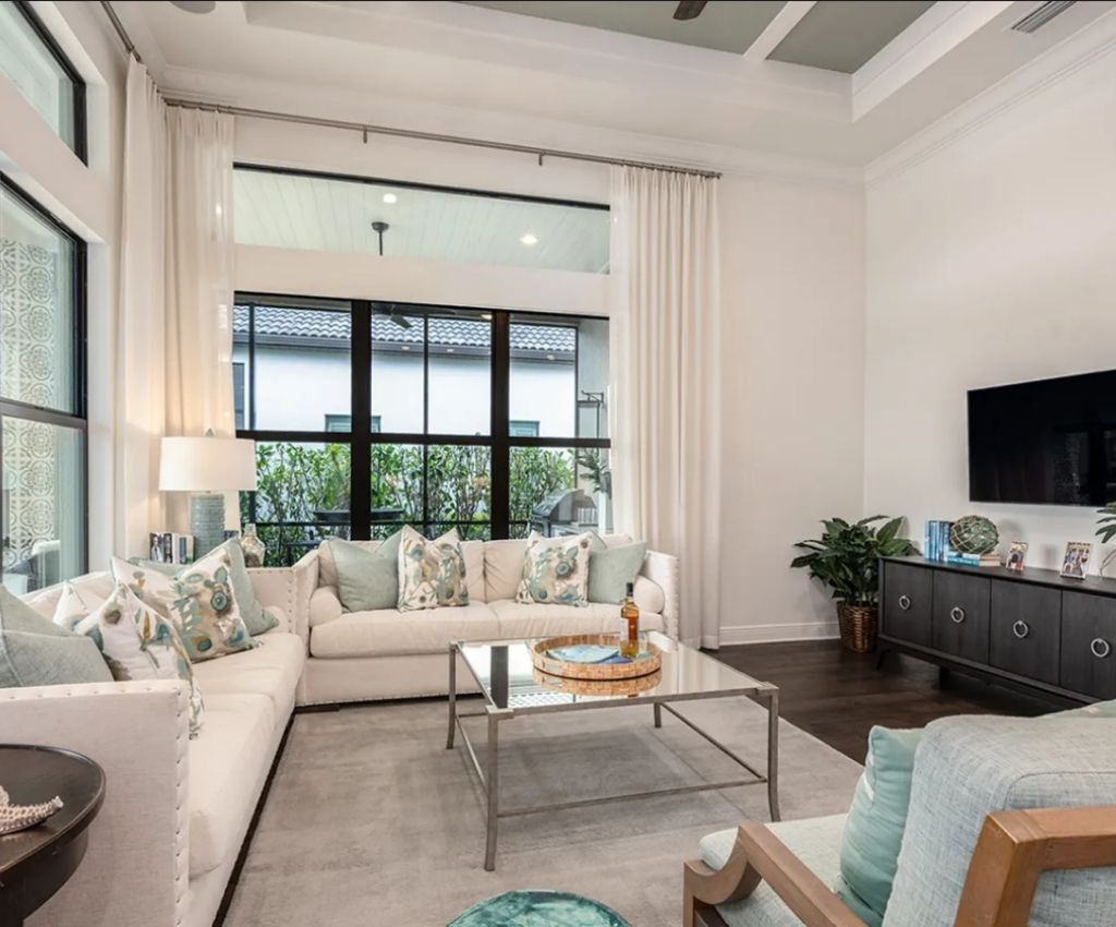 large lounge room with lots of natural light and floor space. L-shaped sofa seating in the back corner, facing the main wall with a mounted TV and cupboard storage underneath it. glass table sits in the center for convenience and colourful accessorise are around the room to create a welcoming atmopsphere