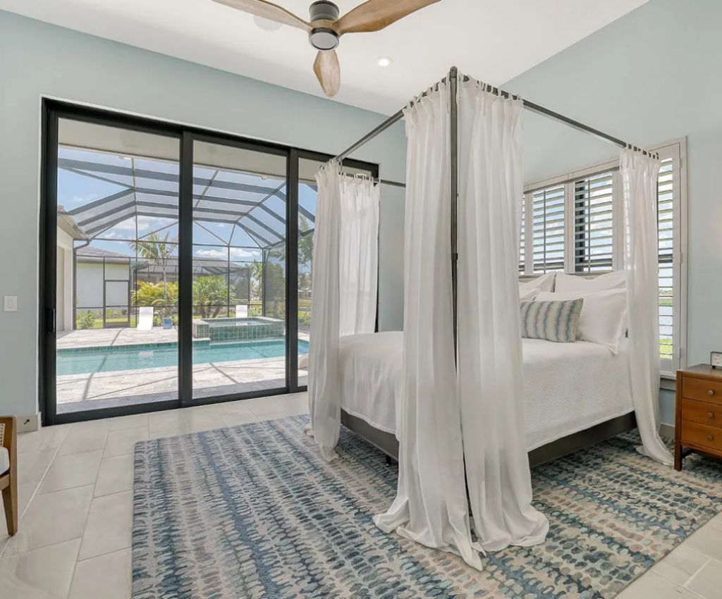 classy bedroom suite with four poster bed with white voile drapes around it sits beside dark wood framed sliding glass doors leading out to the glass roofed lanai area with a pool.