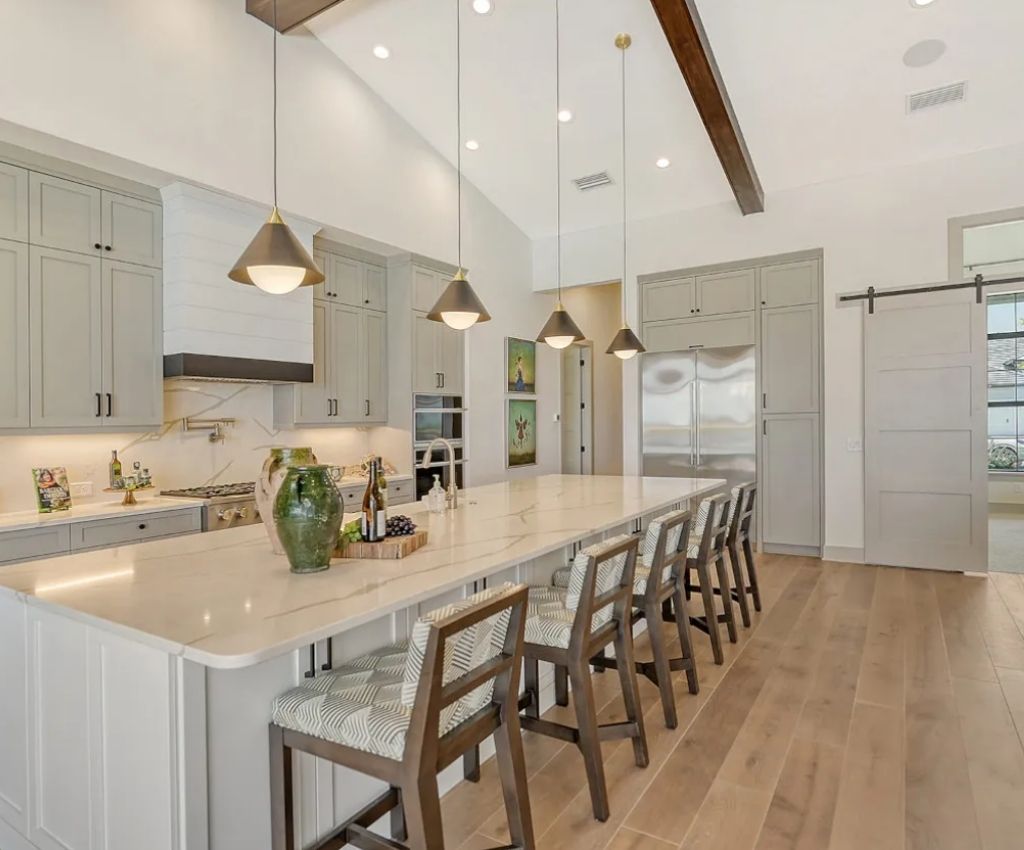 long view of the main kitchenarea - gloss cupboards line the wall, a matching island/breakfast bar separates the kitchen from the living area, and dark wooden bar chairs sit at it. Pendant lights hang down over the island.