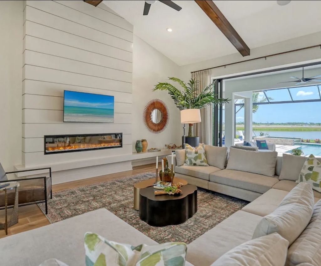 inside the main living area of the palm bay 2 home floor plan: large window on the right with lake views, a tall plant in the corner makes it feel homely, L-shaped seating is arranged around the main wall which has a fire built into it.