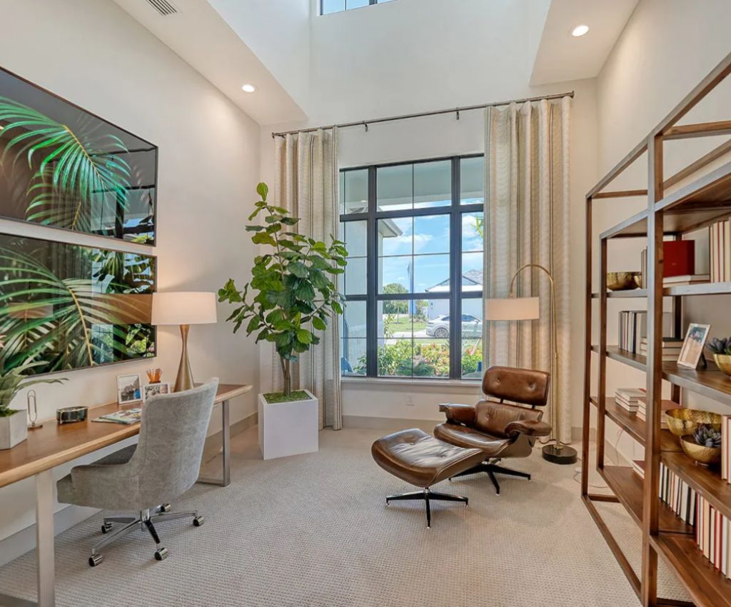 a stylish home office space is lit with tall windows and natural light and atmospheric wall lamps. A desk runs down one side fo the room, freestanding shelves down the other, and a leather recliner chair and footstool sit by the window at the end of the room.