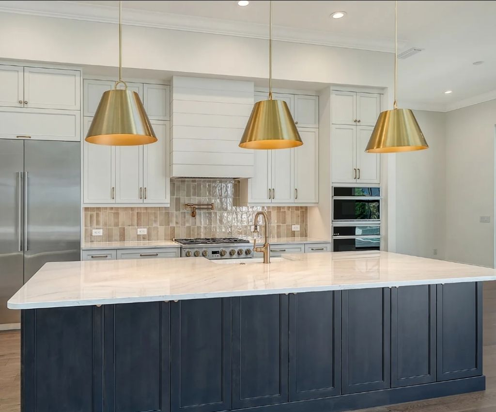 dark blue kitchen island with sleek white counter top, three gold hanging lights sit above it, and white cupboards line the wall behind it