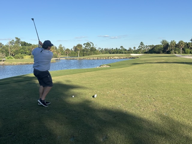 sun shines down on lush green grass at esplanade golf course, a long lake stretches on the left side, and Josh swings his club to tee off.