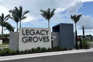 large grey stone sign at the side of the road with "legacy groves" on it, surrounded by palm trees